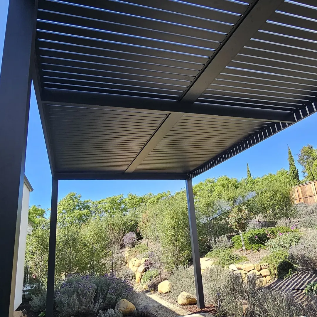 Underside view of a black aluminum slat patio cover with center framing over a landscaped backyard