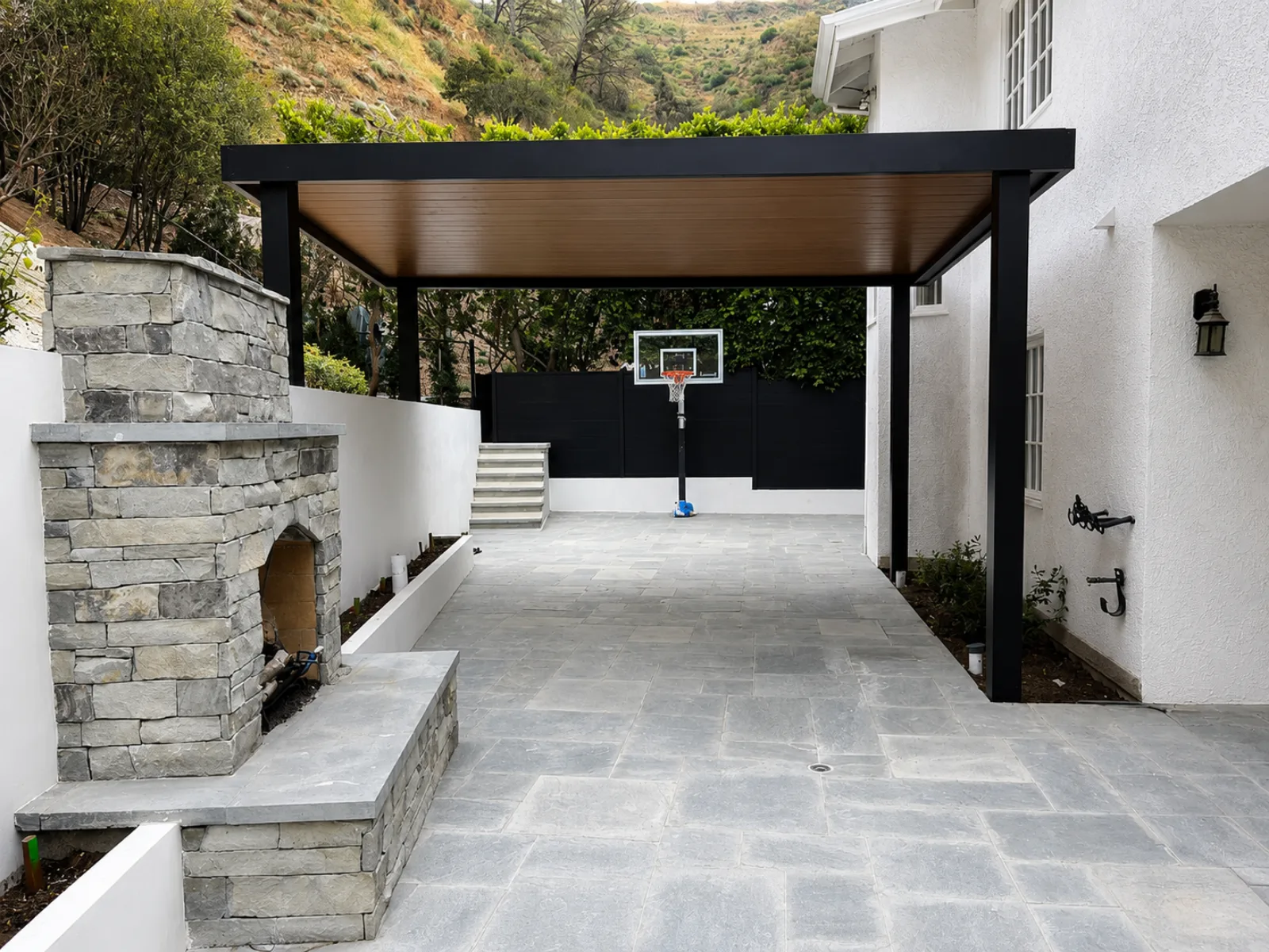 Freestanding black aluminum patio cover with a wood-look insulated roof beside a backyard basketball court